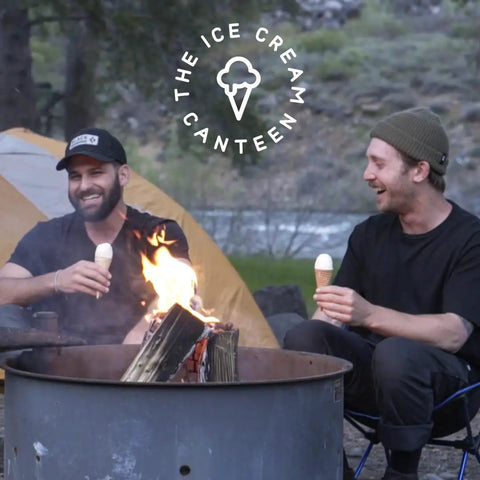 Two friends enjoying ice cream cones near a campfire, surrounded by nature, with a tent in the background.