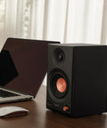 Black MR3 Powered Studio Monitors With Hi-Res Audio Certification And Bluetooth 5.4 next to a laptop and mouse on a desk.