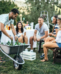 People at an outdoor gathering with a wheelbarrow filled with beverages and ice, chairs, and decorations in a garden setting.