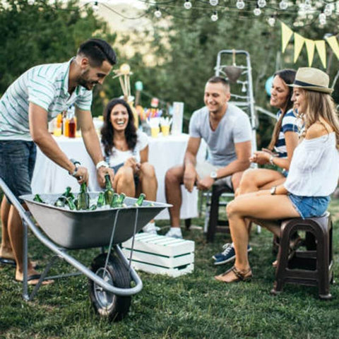 People at an outdoor gathering with a wheelbarrow filled with beverages and ice, chairs, and decorations in a garden setting.