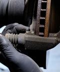 Close-up of gloved hand applying AGS SIL-Glyde silicone brake lubricant to a rusty caliper.