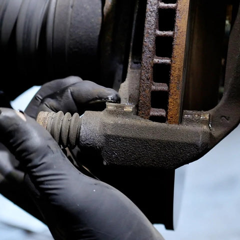 Close-up of gloved hand applying AGS SIL-Glyde silicone brake lubricant to a rusty caliper.