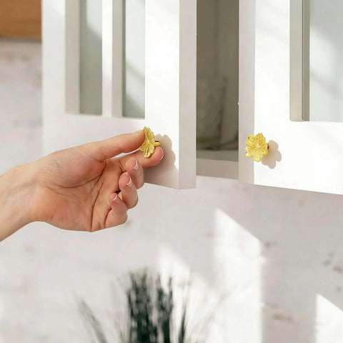 A hand reaching for a vintage gold flower cabinet knob on white cabinetry, showcasing its floral design, part of the eight-piece hardware set. Kickstart Your Home Refresh With Vintage Gold Flower Cabinet Knobs And Brass Drawer Pulls An Eight Piece Hardware Set Delivering Next Level Swag To Every Kitchen Or Bedroom.