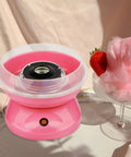Pink cotton candy machine with white guard cover enclosure next to a cotton candy serving and strawberry on a glass bowl