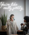 Two individuals talking under a 'You're like really pretty' neon sign, indoor plant and table visible.