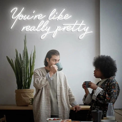 Two individuals talking under a 'You're like really pretty' neon sign, indoor plant and table visible.