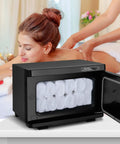 Pro 8L Towel Warmer For Salon And Spa in black, filled with rolled towels, next to a woman receiving a back massage in a spa setting.