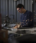 Worker using Heavy Duty Sixty Gallon Two Stage Air Compressor during metal grinding, sparks flying in workshop.