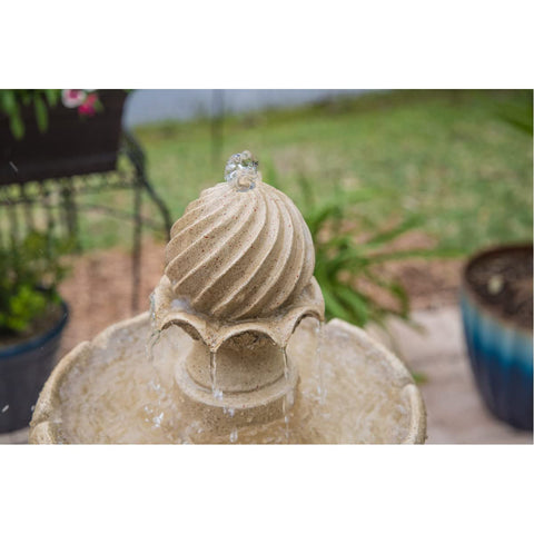 Outdoor stone fountain with water flowing, spiral finial detail, surrounded by green plants and pottery in a garden setting.