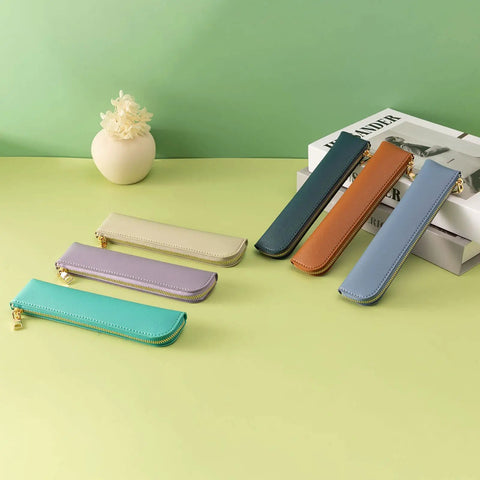 Assorted slim leather pencil cases with zippers on a green surface, showcasing different colors including purple, blue, and brown.