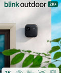 Level Up Your Home Security Game With This Black Outdoor Wireless Camera mounted on a wall with greenery.
