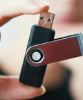 Close-up of a hand holding a swiveling USB flash drive with a metallic finish against a blurred background.