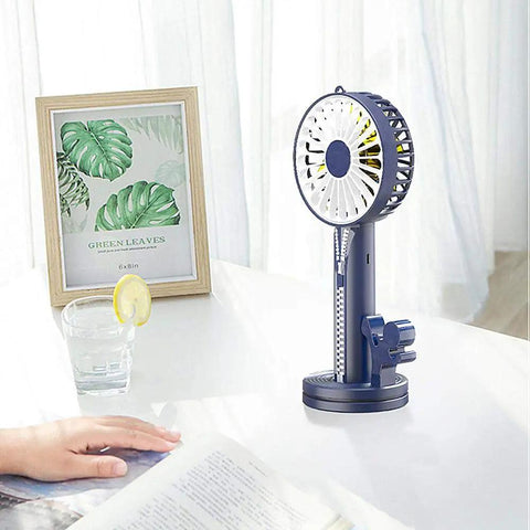 Portable desk fan on a table with a glass of water, framed leaf print, and open book