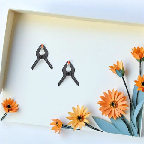 Top Tier Heavy Duty Spring Clamps Set For Woodworking And Photography Backdrops, Elastic Plastic Clips With Compact Design Great For Any Creative Project. Two black spring clamps on white board with paper flowers