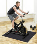 Man exercising on a stationary bike placed on a black mat in a home gym setting with wooden floor and nearby dumbbells.