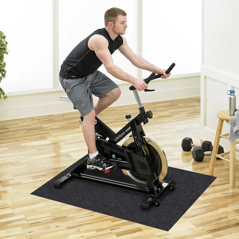 Man exercising on a stationary bike placed on a black mat in a home gym setting with wooden floor and nearby dumbbells.
