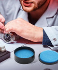 Watchmaker repairing watch near Pro Grade Watch Gasket Grease Silicone Lubricant For O-Rings And Case Backs, visible in black and blue.