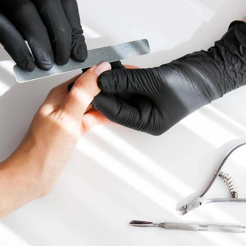 Black disposable vinyl gloves worn on a gloved hand, with a metal nail file and manicure tools on a white surface.