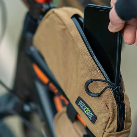 Person inserting phone into Toprock Frame Bag For Road And Gravel Cycling Water Resistant Cordura 1000D With Durable YKK Aquaguard Zipper Top Tube Frame Storage For Daily Essentials And Bikepacking Coyote Color 1 Liter attached to bike