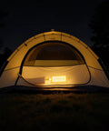 A glowing yellow camping tent illuminated from inside at night, set up on a grassy field under a dark sky.