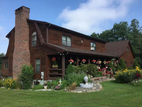 Top Shelf Heavy Duty Oil Based Wood Stain For Exterior Decks Fences Siding Logs Featuring Knockout UV Shielding And Deep Soak Water Repellent Formula In Easy Roll Semi Transparent Rustic Brown Covers Large Patios applied to a log cabin home with flower gardens.