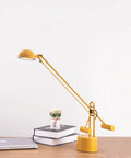 Yellow desk lamp with counter-balance arm on a wooden desk beside a laptop and books, modern office task lighting.