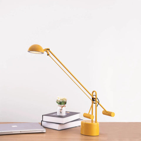 Yellow desk lamp with counter-balance arm on a wooden desk beside a laptop and books, modern office task lighting.