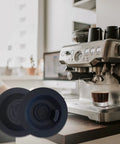 Two blue 54mm espresso backflush cleaning discs, 1 pack, on a kitchen counter beside a stainless espresso machine.