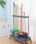 Stainless Steel Broom And Mop Organizer Rack With Five Racks And Six Hooks, holding various cleaning tools, floor mounted, in a room.