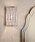 Wall-mounted light fixture with textured glass and metal design next to a bed headboard.