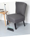 Grime Crushing Heavy Duty Broom With 34 Inch Long Handle For Sweeping Concrete Driveway Garage Patio And Indoor Floors No Sweat Household Cleaner With Convenient Storage Hook next to a chair on a grey floor.