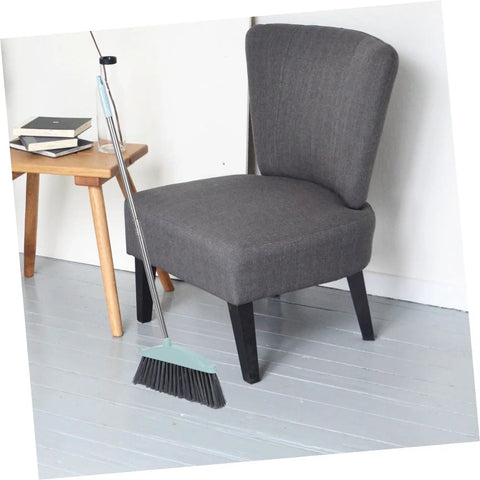 Grime Crushing Heavy Duty Broom With 34 Inch Long Handle For Sweeping Concrete Driveway Garage Patio And Indoor Floors No Sweat Household Cleaner With Convenient Storage Hook next to a chair on a grey floor.