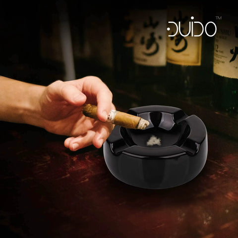 Hand holding cigar over Large Eight Point Five Inch Black Glossy Ceramic Cigar Ashtray Kit With Stainless Steel Cutter