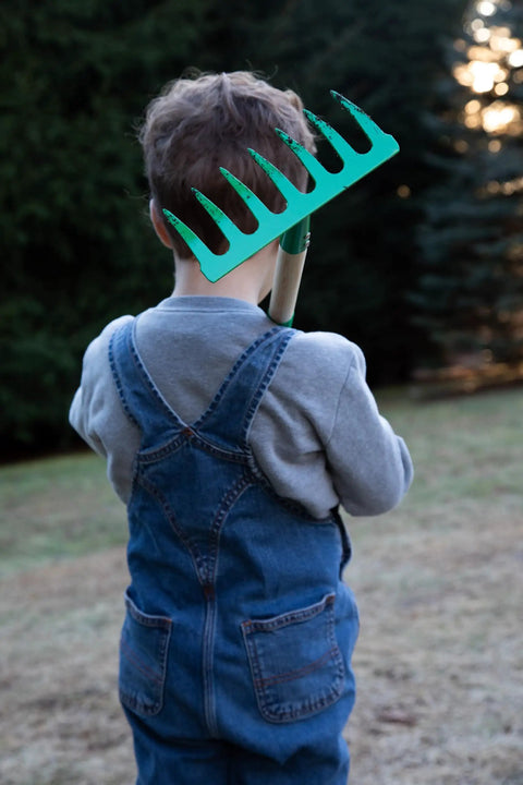 Child holding Kids Garden Tool Set 4 Piece Rake Spade Hoe And Leaf Rake With Real Wood Handles And Steel Heads, outdoor setting.