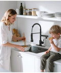 RNDIOZD LED Kitchen Faucet with Pull Down Sprayer, 2-Spout, Matte Black in modern kitchen with woman and child.