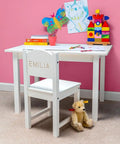 Children's white desk and chair with colorful art supplies and toys against a pink wall, featuring 'Emilia' personalization on the chair.