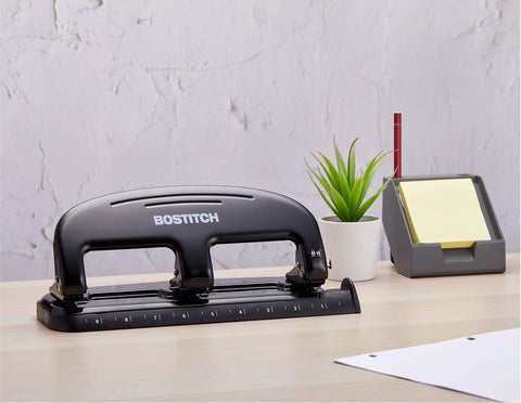 Bostitch 20-sheet 3-hole punch, metal construction, black; desk scene with plant and note organizer.
