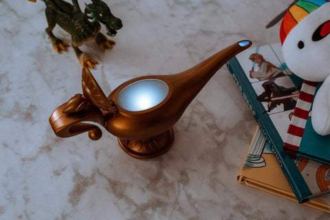 Aladdin Genie Lamp LED Mood Light 10 Inch Blue And White Mood Lighting Figure Collectible Genie Lamp Decor Epic Glow Night Light For Fans And Collectors beside books and toy dinosaur on marble surface.