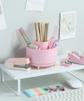 Pink Desk Accessories Set With Rotating Pen Holder, Stapler, Staple Remover, Tape Dispenser And Refills on a white shelf.