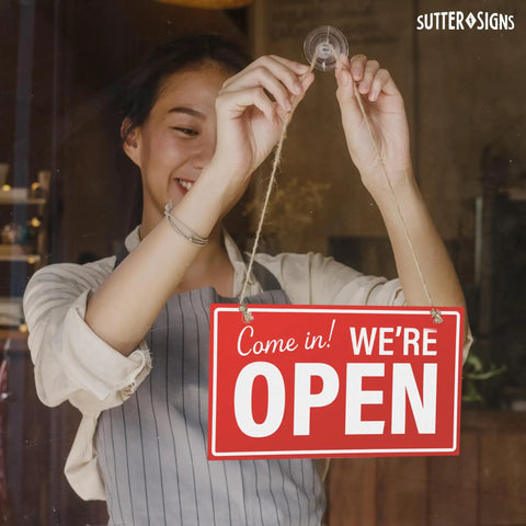Person hanging Open And Closed Sign For Biz 10 By 6 Inches Double Sided Plastic Sign With Twine And Suction Cup Hook For Door Or Window Easy To Read Business Hours Black And Red on glass door