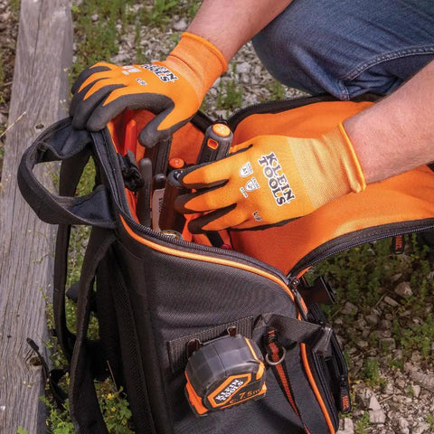 Person wearing orange knit dipped cut resistant work gloves with nitrile coated palms, handling tools from a black bag outdoors.
