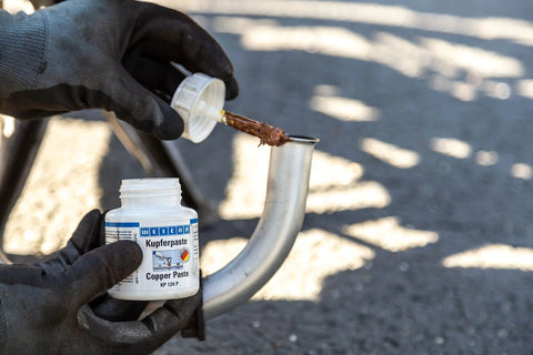 WEICON Copper Paste bottle (4.2 oz/120 g) being applied to a curved metal joint by gloved hands.