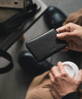 Person holding Ultra Slim Minimalist Card Holder With RFID Blocking Leather Front Pocket Wallet in black, above table and coffee cup.