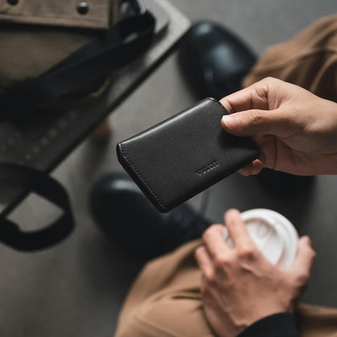 Person holding Ultra Slim Minimalist Card Holder With RFID Blocking Leather Front Pocket Wallet in black, above table and coffee cup.