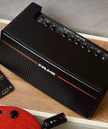 30W Wireless Modeling Guitar Amplifier With Built In IRs Dual 4 Inch Coaxial Speakers on a wooden table with remote and guitar.