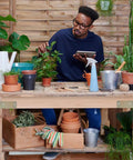 Person in blue shirt working with plants at a wooden workbench in a garden setting, surrounded by pots and gardening tools.