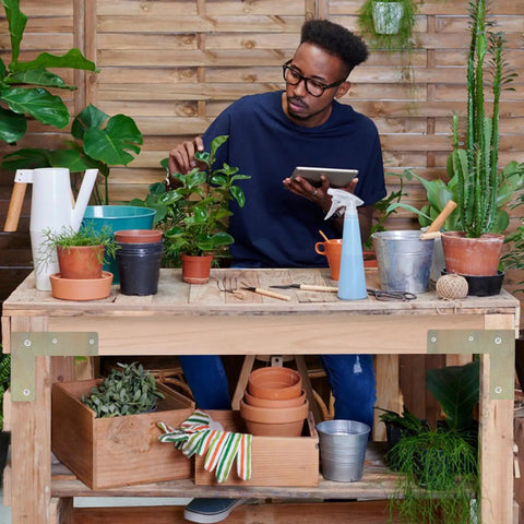 Person in blue shirt working with plants at a wooden workbench in a garden setting, surrounded by pots and gardening tools.