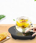 Solid Daily Driver One Button Cup Warmer Heating Plate With Lightning Fast Five Second Heat Up And Zero Leak Constant Temperature For Home Coffee, with glass mug on a black heating plate, showing a hand pressing the on/off button.