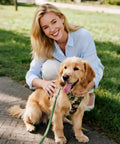 Woman kneeling and smiling with a golden retriever wearing a camouflage harness outdoors on a sunny day.