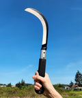 Hand holding K310 Brush Clearing Sickle With 9 Inch SK5 Japanese High Carbon Steel Blade And 9 Inch Aluminum Handle in clear sky.
